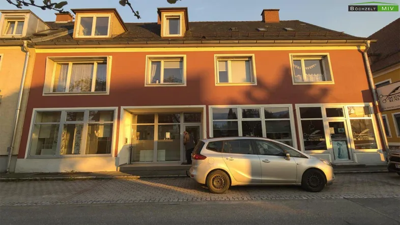 Rote Fassade eines Gebäudes mit mehreren Fenstern und einem geparkten Auto davor auf der Straße.