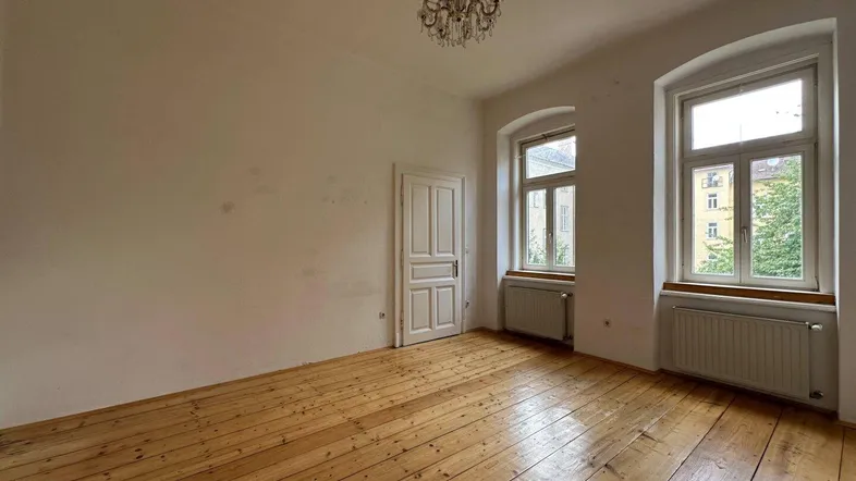 Geräumiges Zimmer mit hohen Decken, Dielenboden und zwei großen Fenstern, die viel Licht hereinlassen.