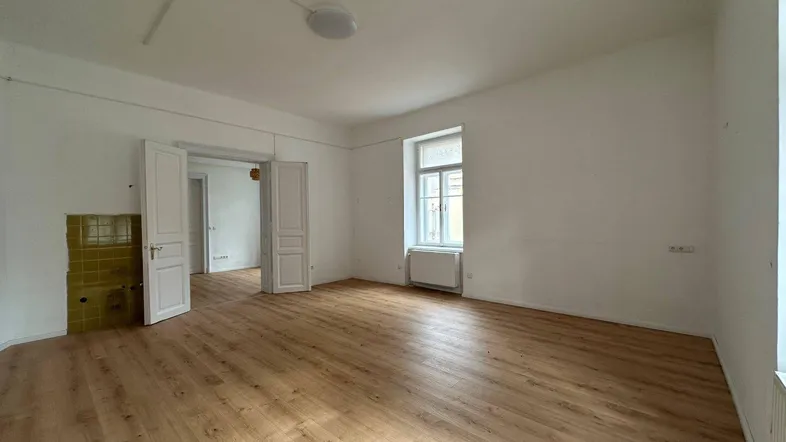 Geräumiges Zimmer mit hellem Laminatboden, hohen Decken und einem traditionellen Kachelofen.
