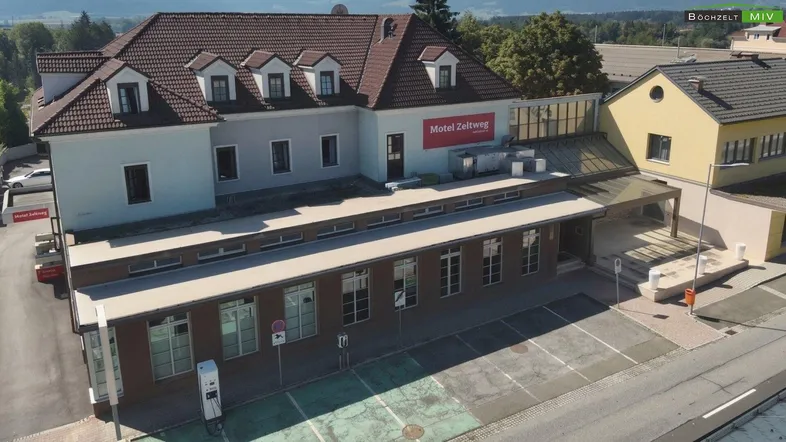 Luftaufnahme des Gebäudes mit Parkplätzen und Ladesäulen, umgeben von Bäumen und anderen Gebäuden.