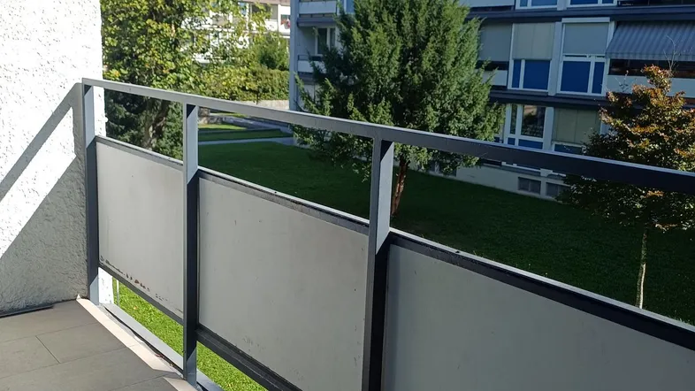 Balkon mit Blick auf eine grüne Rasenfläche und umliegende Gebäude unter blauem Himmel.