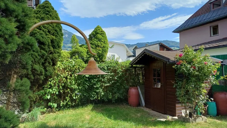 Gartenbereich mit einem Holzschuppen, Rosen und grünen Pflanzen unter blauem Himmel.