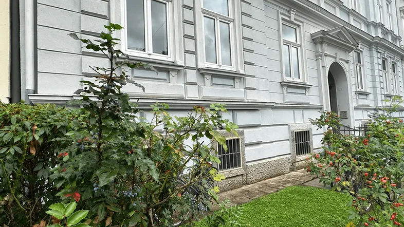 Graue Fassade eines Altbaus mit weißen Fenstern und gepflegtem Vorgarten.
