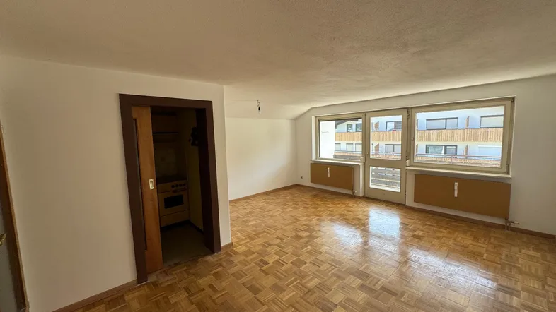 Helles Wohnzimmer mit Parkettboden, großen Fenstern und Zugang zu einer kleinen Küche.