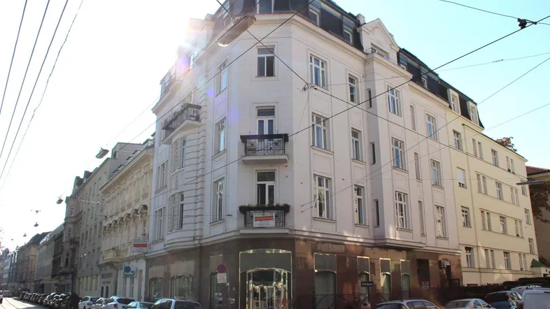 Eckgebäude mit klassischer Architektur, Balkonen und sonniger Fassade an einer belebten Straße.