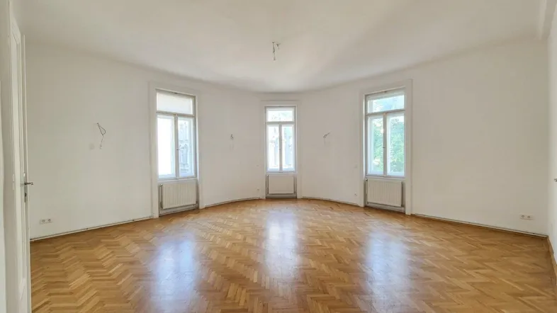 Helles, leeres Zimmer mit Fischgrätparkett und drei hohen Fenstern, bereit zur individuellen Gestaltung.