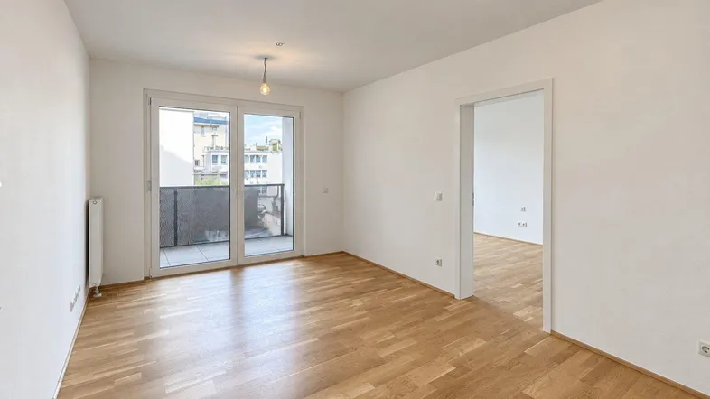 Helles, leeres Wohnzimmer mit Parkettboden und Zugang zu einem Balkon.