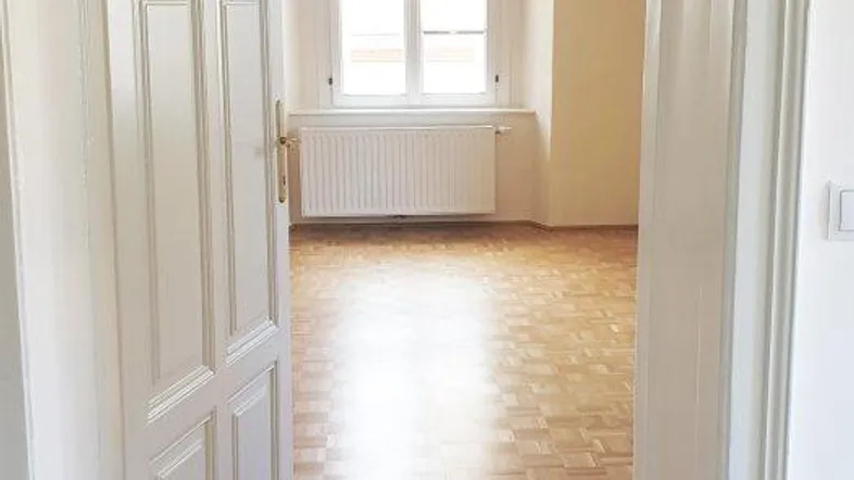 Blick durch eine offene weiße Tür in einen hellen Raum mit Fischgrätparkett und Fenster.