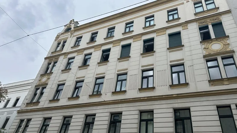 Historische Fassade eines Altbaus mit vielen Fenstern und dekorativen Elementen unter bewölktem Himmel.