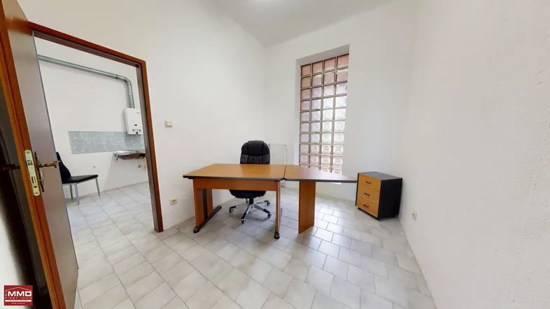 Helles Büro mit Schreibtisch, Bürostuhl und einem Fenster aus Glasbausteinen, angrenzend an einen Raum mit Spülbecken.