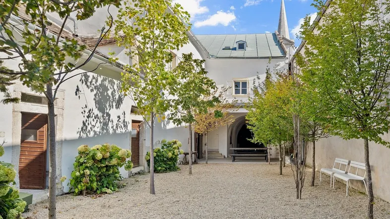 Historischer Innenhof mit Kiesboden, jungen Bäumen und Sitzbänken, umgeben von Gebäuden.
