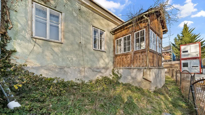 Altes Haus mit verputzter Fassade und einem hölzernen Anbau, umgeben von wilder Vegetation.