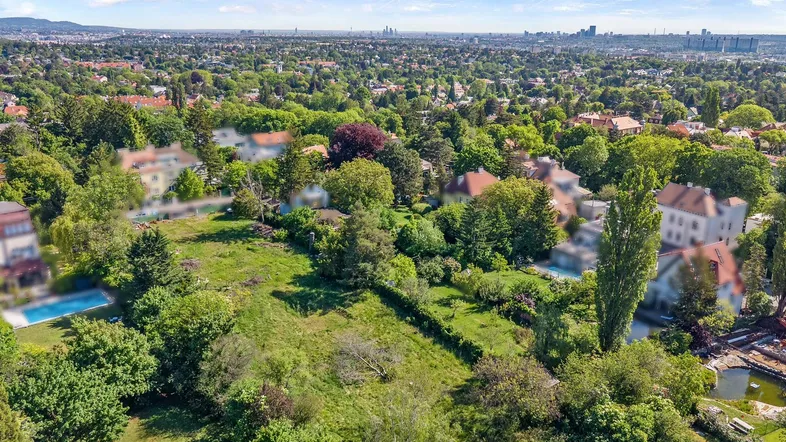 Luftaufnahme eines weitläufigen grünen Grundstücks mit umliegenden Häusern und Stadtblick in der Ferne.