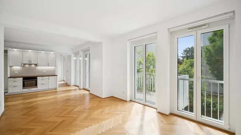 Helles Wohn- und Esszimmer mit Fischgrätparkett, offener Küche und Zugang zum Balkon.