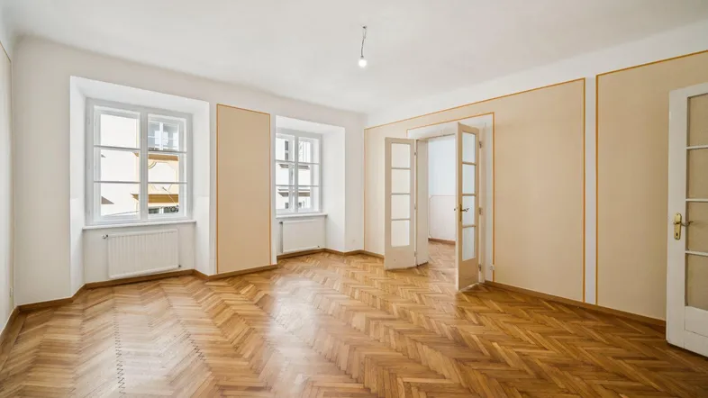 Helles Zimmer mit Fischgrätparkett, zwei Fenstern und Doppeltüren, ideal als Wohn- oder Schlafzimmer.