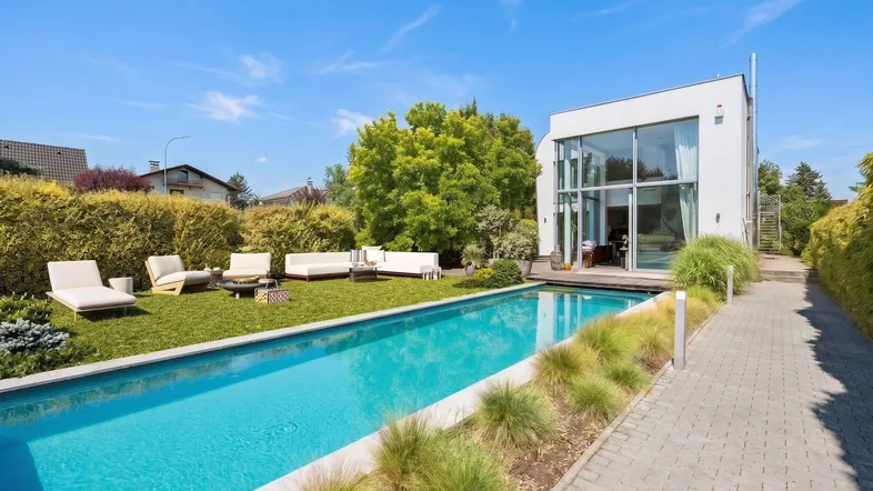 Modernes Haus mit großem Garten, langem Pool und gemütlicher Outdoor-Sitzgruppe unter blauem Himmel.