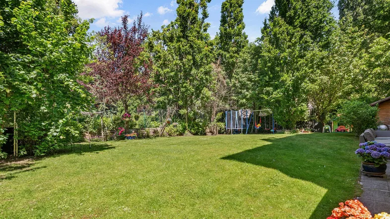 Großer, gepflegter Garten mit weitläufiger Rasenfläche, hohen Bäumen und Spielgeräten für Kinder.