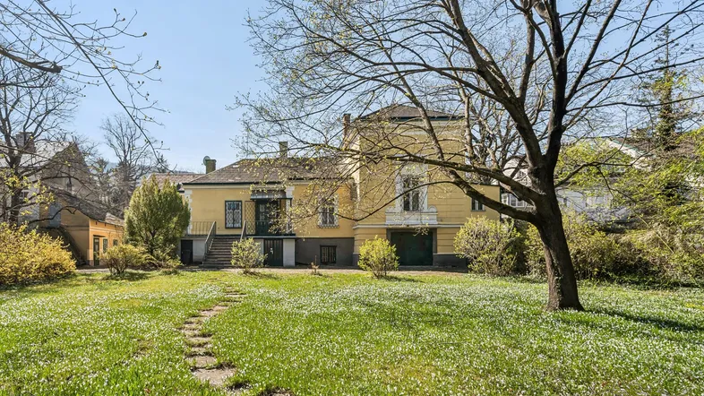 Gelbe Villa mit großem Garten und altem Baumbestand unter blauem Himmel.
