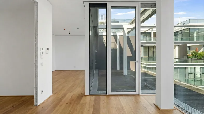 Heller Innenraum mit Holzboden und großen Glasfronten, die zu einer Terrasse führen.