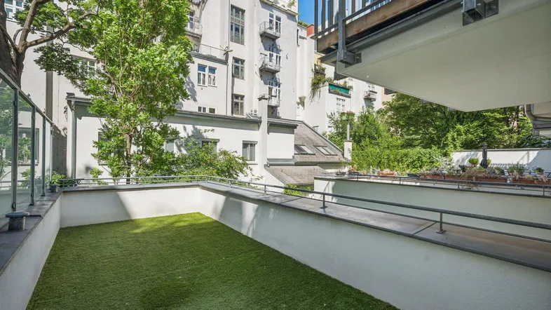 Große Terrasse mit Kunstrasen, Glasgeländer und Blick auf die umliegenden Gebäude und Bäume.