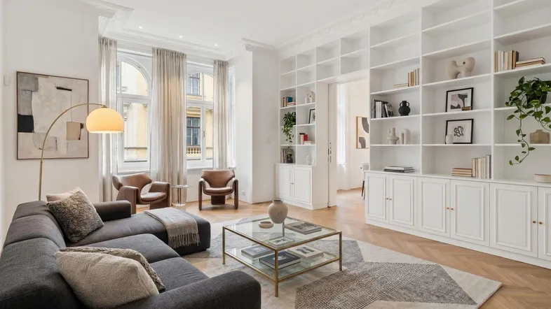 Helles Wohnzimmer mit modernem Sofa, Bücherregalen und hohen Fenstern, die viel Tageslicht hereinlassen.