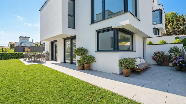 Moderne weiße Villa mit gepflegtem Rasen und sonniger Terrasse mit Sitzgelegenheiten.