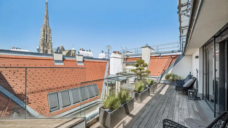 Geräumige Dachterrasse mit Holzboden, Glasgeländer und Panoramablick über die Stadt und den Stephansdom.