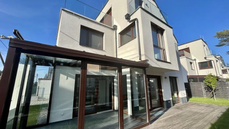 Modernes Haus mit hellem Wintergarten und Holzterrasse, umgeben von Grünflächen.