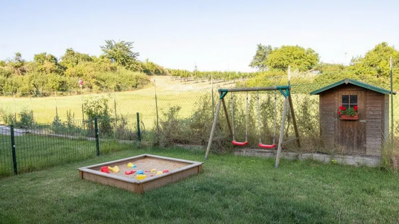 Spielbereich im Garten mit Sandkasten, Schaukel und Gartenhaus, umgeben von grüner Landschaft.