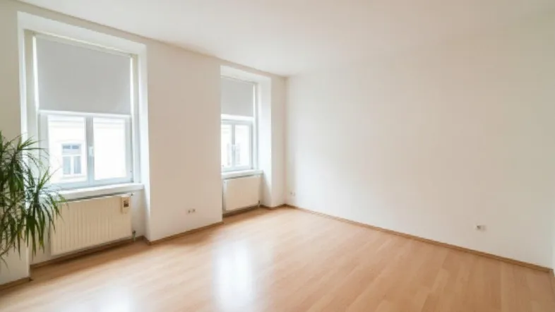 Helles, leeres Zimmer mit zwei Fenstern, hellem Parkettboden und einer Pflanze.