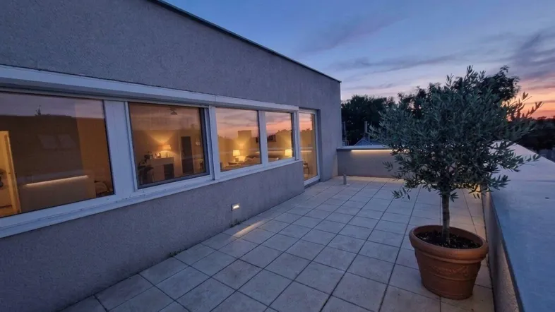 Geräumige Dachterrasse mit Fliesenboden, Topfpflanze und stimmungsvoller Abendbeleuchtung bei Sonnenuntergang.