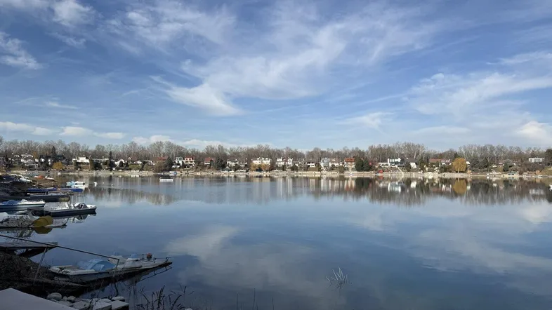 Weitläufiger Blick über den See mit Häusern am gegenüberliegenden Ufer unter blauem Himmel.