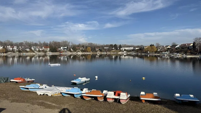 Blick auf einen See mit bunten Tretbooten am Ufer und Häusern im Hintergrund unter blauem Himmel.