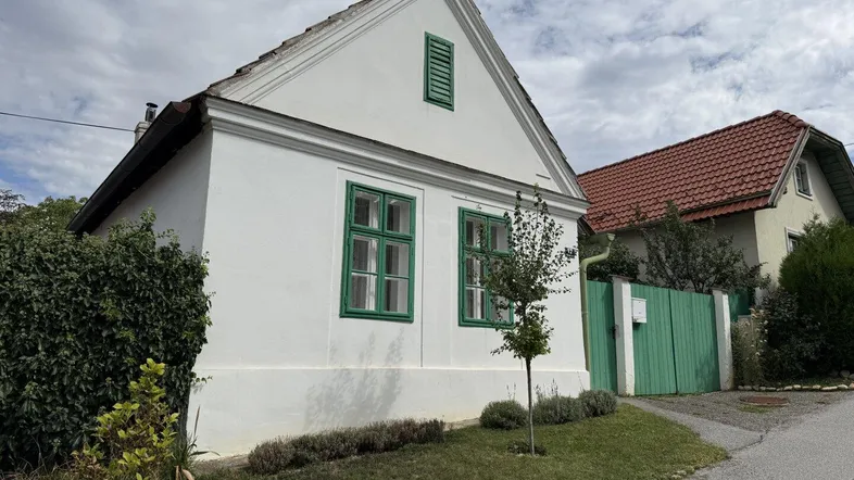Weißes Haus mit grün gestrichenen Fenstern und Giebeldach unter bewölktem Himmel.