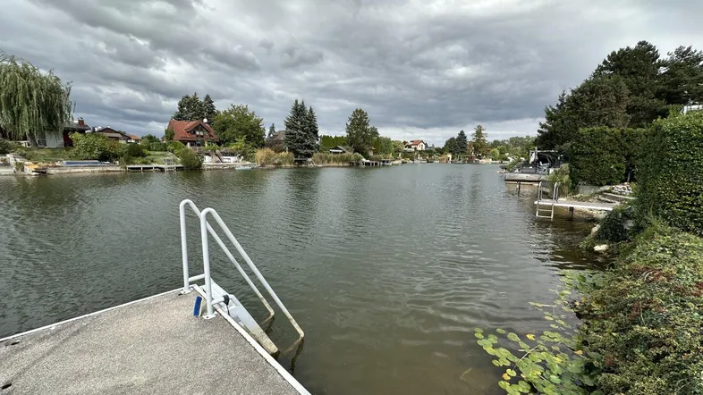 Blick auf einen ruhigen Kanal mit Anlegestelle und Häusern am Ufer unter bewölktem Himmel.