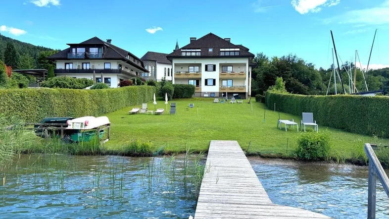 Seeblick auf zwei Mehrfamilienhäuser mit gepflegtem Rasen, Hecken und einem privaten Steg am Wörthersee.