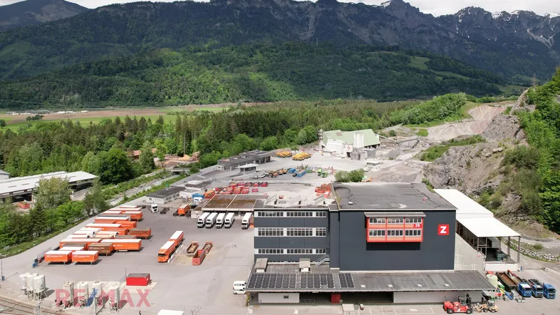 Luftaufnahme eines Industriegebiets mit Lagerhallen, LKW-Parkplätzen und Bergen im Hintergrund unter blauem Himmel.