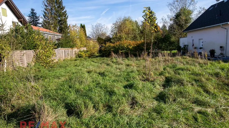 Unbebautes Grundstück mit Wiese und umgebenden Häusern unter blauem Himmel.
