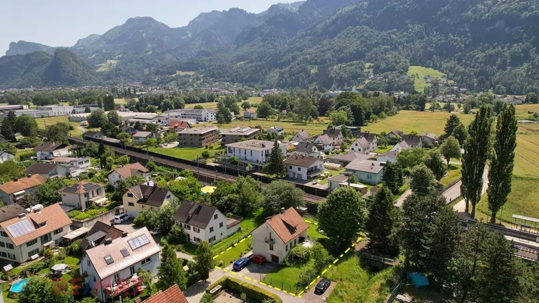 Luftaufnahme des Hauses mit umliegender Wohngegend, viel Grün und Bergpanorama im Hintergrund.