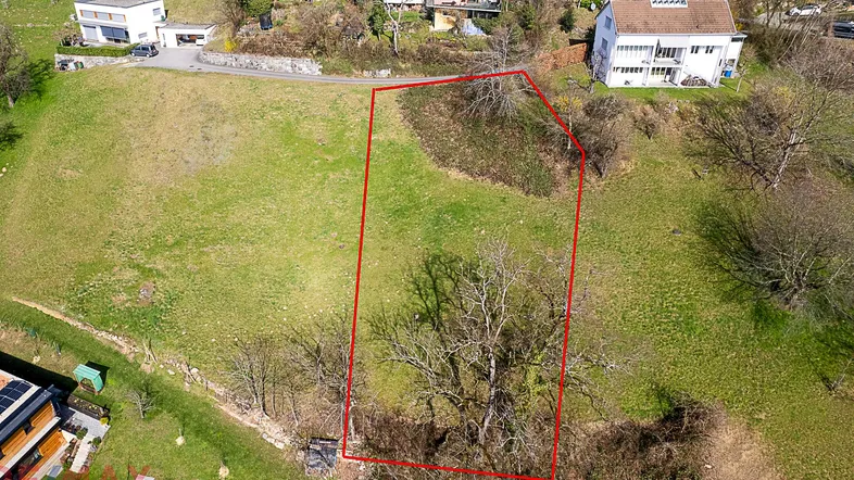 Luftaufnahme eines Baugrundstücks mit roter Umrandung, umgeben von grünen Hügeln und Wohnhäusern.