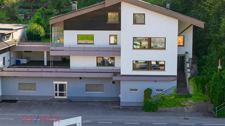 Mehrstöckiges Gebäude mit weißer und grauer Fassade, umgeben von grüner Vegetation und Balkonen.
