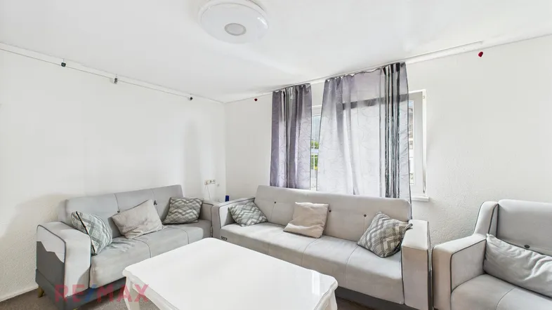 Helles Wohnzimmer mit bequemen grauen Sofas, einem weißen Couchtisch und einem Fenster mit Vorhängen.