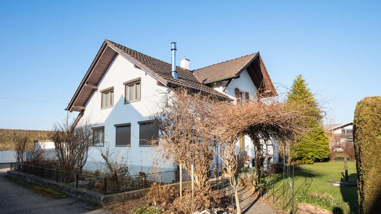 Weißes Einfamilienhaus mit Satteldach, Rollläden und einem kleinen Vorgarten.
