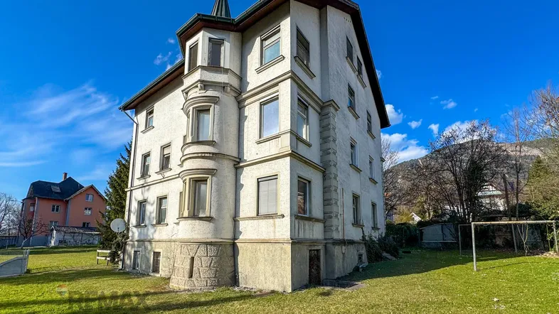 Historisches Gebäude mit markantem Erker und Turmspitze, umgeben von einer grünen Rasenfläche.