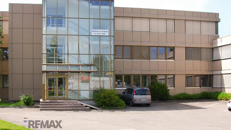 Modernes Bürogebäude mit Glasfassade und Eingangsbereich, umgeben von Parkplätzen und Grünflächen.