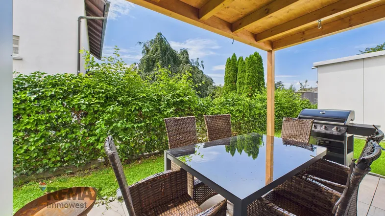 Überdachte Terrasse mit Esstisch, Stühlen und Grill, umgeben von einer grünen Hecke.