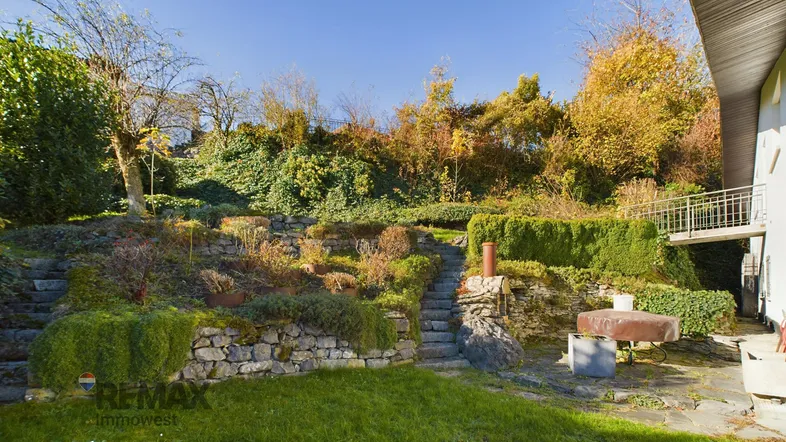 Gestufter Gartenbereich mit Steinmauern, üppiger Vegetation und einem kleinen Balkon am Haus.
