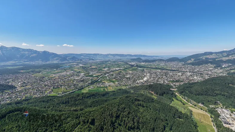 Weitläufige Luftaufnahme einer Stadt im Tal, umgeben von grünen Wäldern und Bergen unter blauem Himmel.