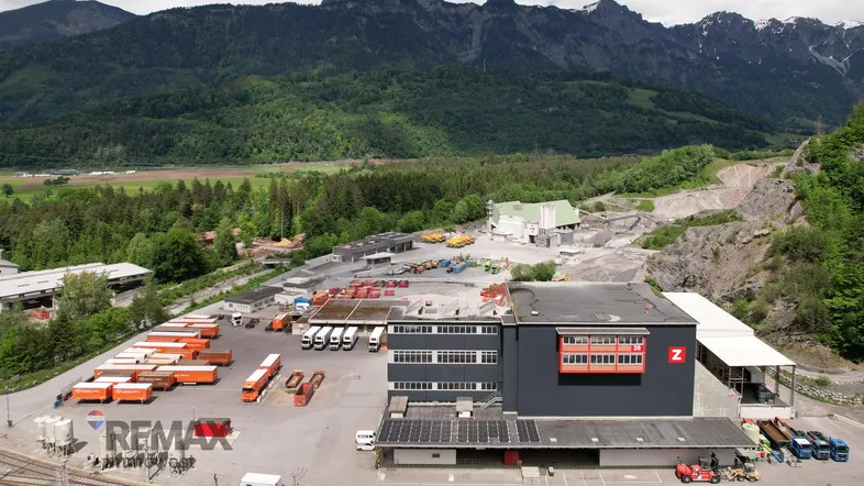 Luftaufnahme eines Industrieareals mit Lagerhallen, Lastwagen und Gleisanlagen vor einer beeindruckenden Bergkulisse.