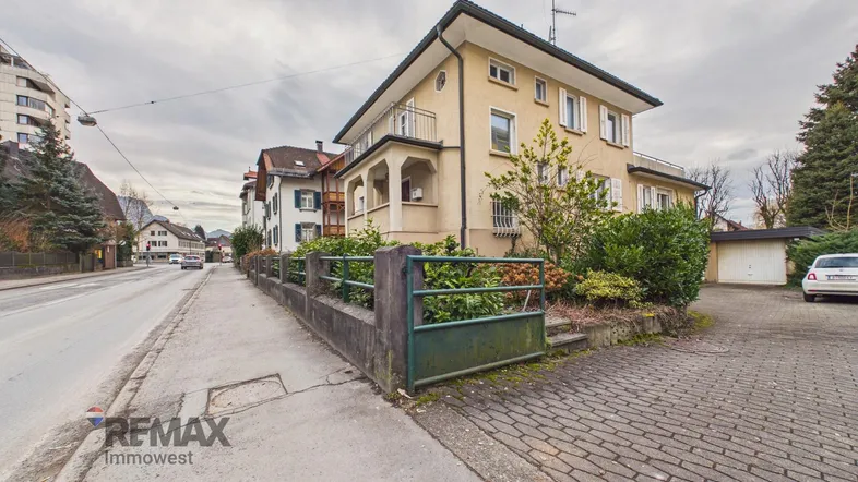 Ansicht des Mehrfamilienhauses von der Straße mit gepflastertem Vorplatz und Hecke.